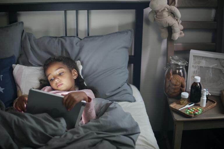 Ребенок лежит в кровати и пользуется планшетом