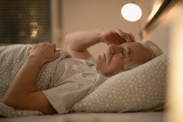 Пожилой мужчина лежит в кровати, держась рукой за голову, потому что страдает бессонницей