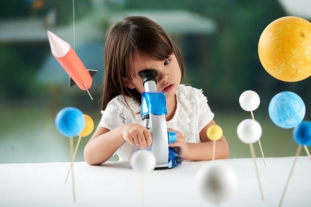 красивая маленькая азиатская девочка смотрит в микроскоп рядом с моделями планет и ракет